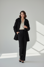Įkelti nuotrauką į galerijos peržiūros programą, A woman stands in a studio wearing a black textured coat over a cream blouse and black trousers, with shadows cast across the floor from a window-like shape.
