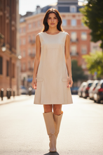 Load image into Gallery viewer, Woman in a beige dress and knee-high boots standing on a city street.