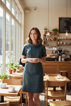Load image into Gallery viewer, Woman standing in a cozy café with wooden furniture and large windows.