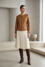 Įkelti nuotrauką į galerijos peržiūros programą, A woman stands in a minimalist concrete room wearing a tan leather jacket, a cream pleated midi skirt, and dark brown knee-high boots.