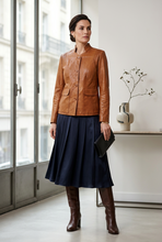 Įkelti nuotrauką į galerijos peržiūros programą, A woman stands indoors near large windows wearing a tan leather jacket, a dark navy midi skirt, and brown knee-high boots, while holding a black clutch.