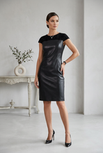 Įkelti nuotrauką į galerijos peržiūros programą, A woman models a black color-block midi dress with a smooth faux-leather finish and a contrasting black velvet V-neckline. She is wearing black patent leather pumps in a minimalist, light gray room with a white console table and a vase in the background.