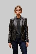 Load image into Gallery viewer, A woman modeling a black leather jacket with a stand collar, worn over a dark gray sweater and dark blue jeans, against a solid gray background.