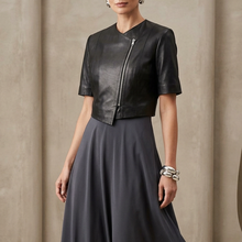 Įkelti nuotrauką į galerijos peržiūros programą, A woman models a black two-piece outfit consisting of a short-sleeve cropped jacket with a front zipper and a matching high-waisted, flowing maxi skirt, set against a neutral textured studio backdrop.