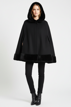 Load image into Gallery viewer, A woman modeling a black hooded cape with plush faux-fur trim along the hood, cuffs, and hem, paired with black pants and black ankle boots, against a light gray studio background.