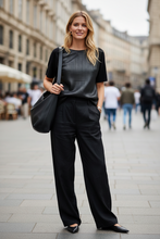 Load image into Gallery viewer, A woman stands on a city street wearing a black short-sleeve ribbed top, black wide-leg trousers, and black pointed flats, while carrying a large black shoulder bag.