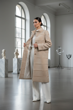 Įkelti nuotrauką į galerijos peržiūros programą, A woman stands in a bright, modern gallery space wearing a long beige textured coat over a white outfit. The setting features arched windows, marble pedestals with sculptures, and a polished gray floor.