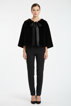 Įkelti nuotrauką į galerijos peržiūros programą, A model stands against a light gray studio background wearing a black velvet cape-style top with a large satin bow at the neckline, black fitted pants, and black heels.