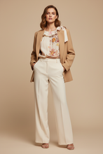 Load image into Gallery viewer, A woman models a stylish outfit consisting of a tan blazer, a cream wide-leg pant, and a floral print blouse with a ribbon detail, set against a solid beige studio background.