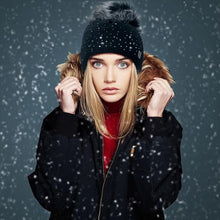 Load image into Gallery viewer, A black knitted cap with a grey artificial fur pom-pom and Swarovski crystals, featuring a small brown label with a brand logo displayed on a woman with stylish outfit.