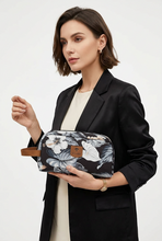 Įkelti nuotrauką į galerijos peržiūros programą, A woman in a black blazer and cream top holds a toiletry bag with a black floral print, white hibiscus flowers, and a brown leather handle. The brand name 'TODD' is visible on a small brown leather patch on the front of the bag.
