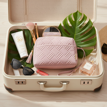 Load image into Gallery viewer, Open suitcase with travel items including a pink makeup bag, sunscreen, and sunglasses on a wooden surface.