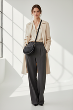 Įkelti nuotrauką į galerijos peržiūros programą, A woman stands in a bright, minimalist white room with window shadows, wearing a beige trench coat over a light blouse, dark gray wide-leg trousers, and black pointed shoes, while carrying a black quilted crescent shoulder bag.