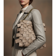 Įkelti nuotrauką į galerijos peržiūros programą, A model wearing a light gray leather jacket and white blouse carries a large woven beige shoulder bag with a gold chain strap against a warm brown studio background.