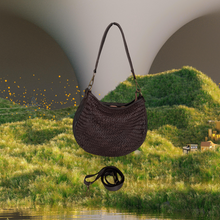 Load image into Gallery viewer, Brown woven handbag with a strap on a grassy hill with a lake and houses in the background