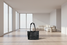 Load image into Gallery viewer, Black tote bag on a light wooden floor with a modern white sofa and large windows in the background