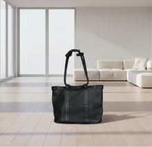 Load image into Gallery viewer, Black tote bag on a light wooden floor with a modern white sofa and large window in the background