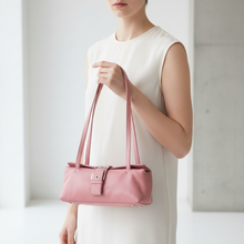 Įkelti nuotrauką į galerijos peržiūros programą, A woman in a sleeveless cream dress holds a structured dusty pink leather shoulder bag with long handles and a front buckle detail, set against a bright, minimalist concrete and white interior background.