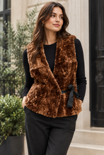 Load image into Gallery viewer, A woman wearing a brown faux fur wrap vest with a black ribbon tie, styled over a black long-sleeve top and dark gray trousers, standing outdoors in front of a building with greenery.