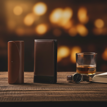 Įkelti nuotrauką į galerijos peržiūros programą, Two cylindrical objects on a wooden surface with a glass of whiskey, watch, and open book against a blurred warm light background.