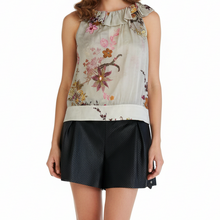 Load image into Gallery viewer, A woman modeling a sleeveless top with a beige base and a colorful floral print, paired with black shorts and black heels, against a plain white background.
