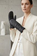Įkelti nuotrauką į galerijos peržiūros programą, A woman in a cream-colored blazer holds up a black leather mitten-style glove, photographed in a minimalist studio setting with a concrete wall background.