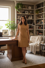 Load image into Gallery viewer, Woman in a beige dress standing in a home office with bookshelves and plants.