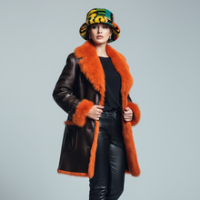 Įkelti nuotrauką į galerijos peržiūros programą, A model wearing a dark brown leather coat with vibrant orange shearling lining, a matching orange shearling collar, a colorful patterned bucket hat, black leather pants, and black high-heeled boots, posed against a light gray studio background.