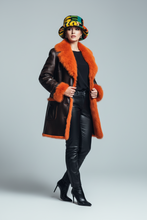 Įkelti nuotrauką į galerijos peržiūros programą, A model wearing a dark brown leather coat with vibrant orange shearling lining, a matching orange shearling collar, a colorful patterned bucket hat, black leather pants, and black high-heeled boots, posed against a light gray studio background.