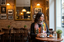 Load image into Gallery viewer, Woman sitting at a table in a cozy cafe with a cup of coffee and pastry.