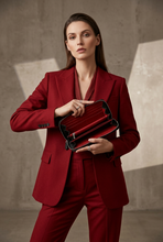 Load image into Gallery viewer, A woman wearing a deep red tailored suit holds open a matching red and black clutch or wallet case against a textured concrete wall with dramatic shadows.