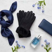 Load image into Gallery viewer, A styled flat-lay featuring a pair of black leather gloves with a dark faux-fur cuff, arranged on a white marble surface alongside a navy blue silk scarf, a navy blue leather wallet, a blue perfume bottle, and small blue flowers.