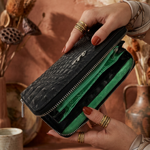 Load image into Gallery viewer, A black embossed leather wallet held open by two hands, showing bright green interior lining and card slots, against a warm rustic background with dried flowers and ceramic vessels.
