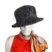 Load image into Gallery viewer, A dark grey astrakhan fur hat displayed on a mannequin wearing a red and orange outfit, with a patterned scarf.