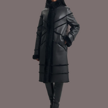 Įkelti nuotrauką į galerijos peržiūros programą, A model wearing a long black faux-leather coat with diagonal stitched panel details and a black fuzzy collar, paired with black pants and boots, against a light gray studio background.