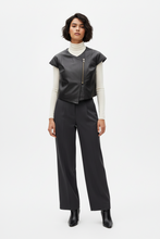 Įkelti nuotrauką į galerijos peržiūros programą, A model stands against a light gray studio background wearing a dark gray faux-leather vest with a gold zipper over a cream turtleneck, dark gray wide-leg trousers, and black ankle boots.