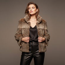 Įkelti nuotrauką į galerijos peržiūros programą, A woman modeling a brown and beige animal-print faux fur jacket over a black satin top and black leather pants, standing against a solid dark gray studio background.