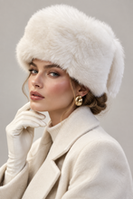 Įkelti nuotrauką į galerijos peržiūros programą, A woman wearing a fluffy cream fur ushanka hat, cream gloves, and a cream coat, with gold statement earrings, photographed against a soft light gray studio background.