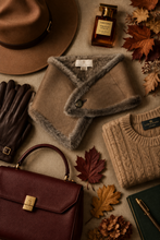 Įkelti nuotrauką į galerijos peržiūros programą, A styled flat-lay of autumn fashion accessories on a linen surface, featuring a taupe suede wrap vest with gray faux-fur trim, a beige ribbed knit sweater, a brown leather handbag, dark brown gloves, a brown wide-brim hat, and a perfume bottle, accented with fall leaves and a pen.