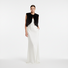 Įkelti nuotrauką į galerijos peržiūros programą, A model stands against a light gray studio background wearing a floor-length white satin gown with a black faux-fur sleeveless vest draped over the shoulders.