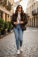 Įkelti nuotrauką į galerijos peržiūros programą, A woman walking down a cobblestone street wearing a brown faux fur jacket, cream turtleneck, light blue jeans, white sneakers, and carrying a black crossbody bag.

