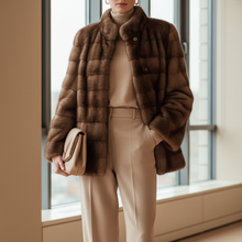 Įkelti nuotrauką į galerijos peržiūros programą, A woman stands in a bright, modern room with large windows, wearing a brown puffer jacket over a beige turtleneck and beige wide-leg trousers, holding a beige clutch.

