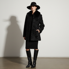 Įkelti nuotrauką į galerijos peržiūros programą, A model stands in a minimalist studio wearing a black textured coat with a plush collar, a black wide-brim hat, a black midi dress, and black knee-high boots, casting a long shadow on the wall.

