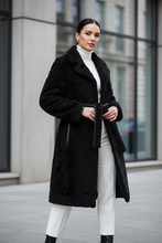 Įkelti nuotrauką į galerijos peržiūros programą, A woman stands outdoors in front of a modern building, wearing a long black textured teddy coat with a tie belt over a white turtleneck and white pants.

