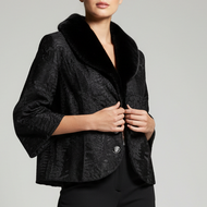 A model stands against a light gray studio background wearing a black textured cropped jacket with a black faux-fur collar, black trousers, and black heeled ankle boots.