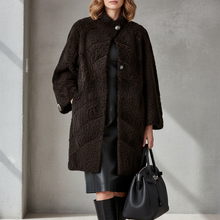 Įkelti nuotrauką į galerijos peržiūros programą, A woman stands in a minimalist studio wearing a dark brown textured teddy coat over a black dress, with black knee-high boots and a black handbag.
