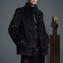 Įkelti nuotrauką į galerijos peržiūros programą, A model wearing a black textured bouclé jacket with a high collar, paired with a black satin dress, set against a dark gray studio background with a gold sculpture.
