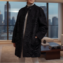 Įkelti nuotrauką į galerijos peržiūros programą, A woman stands in a modern living room wearing a black textured, puffy coat over a light beige turtleneck and white pants. The room features a large window with a city skyline at dusk, a beige sofa, and a wooden coffee table.
