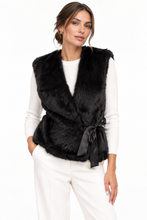 Load image into Gallery viewer, A woman modeling a black faux fur wrap vest with a satin tie belt, worn over a white long-sleeve top and white trousers against a plain white background.
