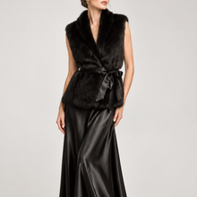 Load image into Gallery viewer, A model wearing a black fur wrap vest with a satin tie belt and a black satin maxi skirt, photographed against a neutral light gray studio background.
